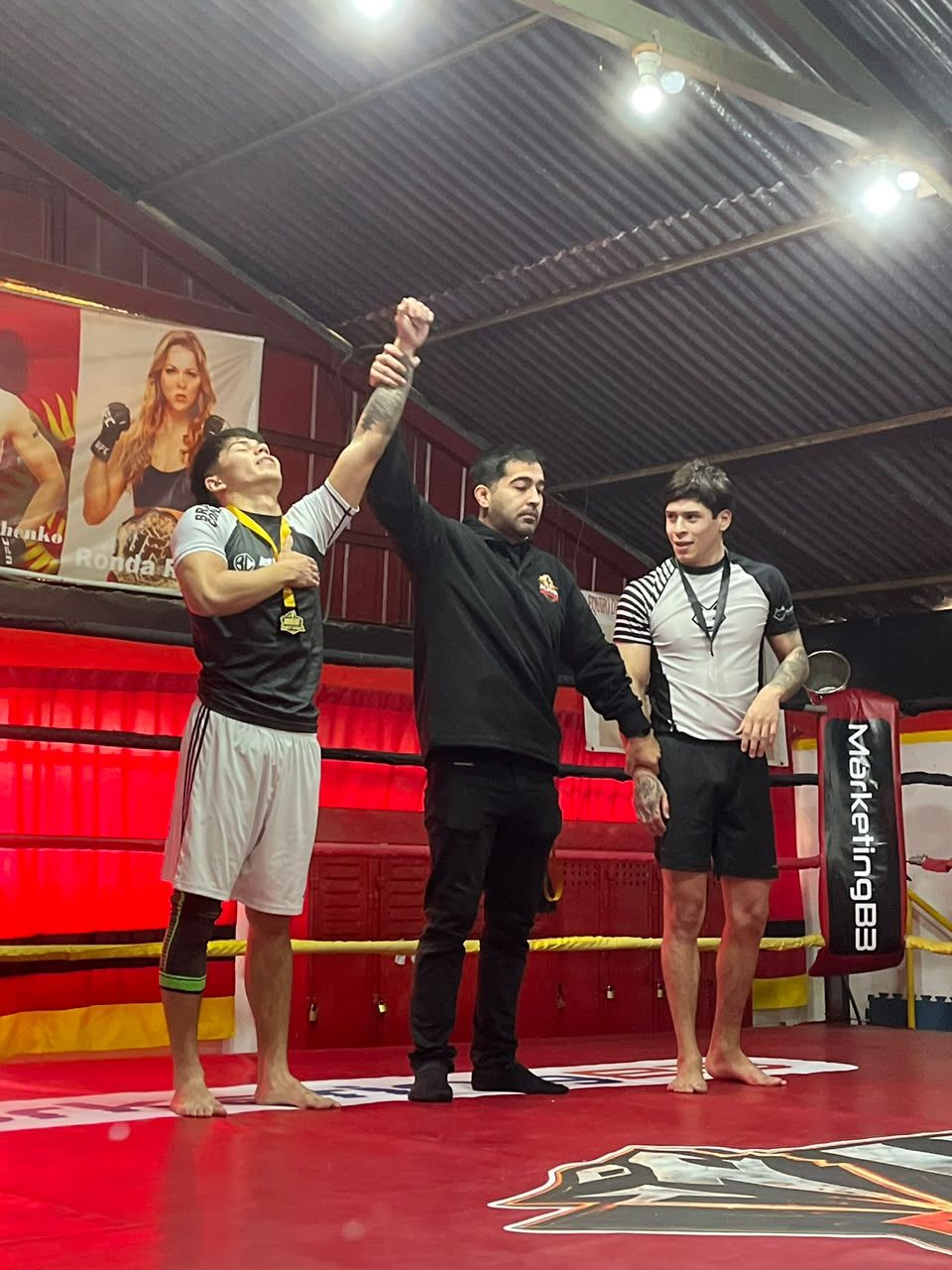 Un alumno con medalla y el brazo levantado junto al árbitro y otro competidor en un ring