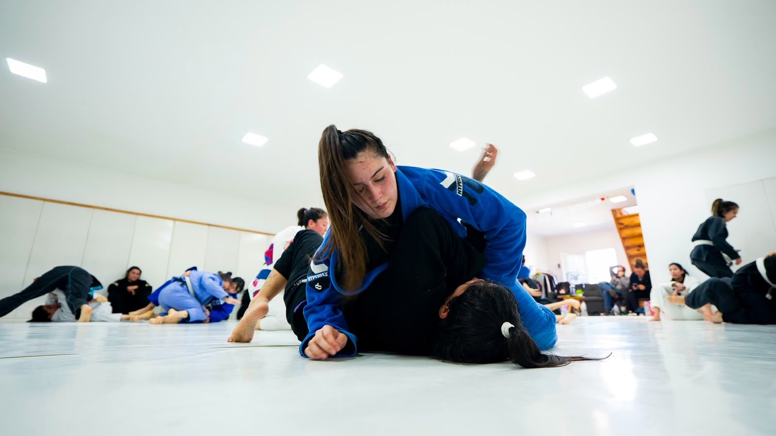 Una persona con kimono azul trabaja una posición sobre su compañera con kimono negro durante una práctica de Jiu-Jitsu